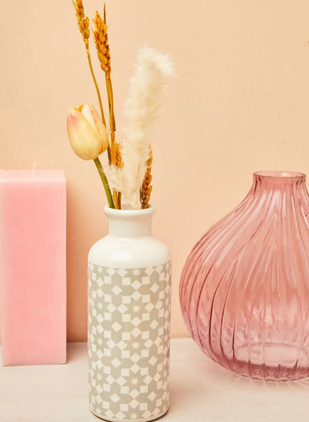 A white ceramic vase with a beige geometric pattern, styled with dried wheat stalks and a pale yellow tulip, set against a soft peach background next to a pink candle and a ribbed pink glass vase.